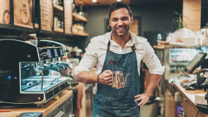Young man working in a coffee shop. He wears a blue apron and holding a metal coffee pot. Heis standing next to a large coffee machine and behind the counter. He is looking and smiling at the camera.
