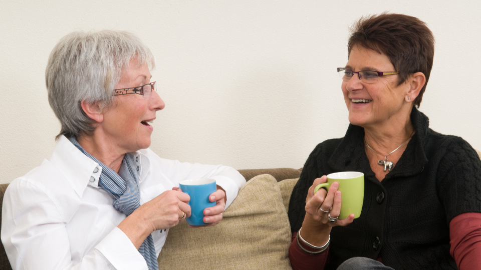 Two women sitting on a sofa talking animatedly to each other while holding a cup in each hand.
