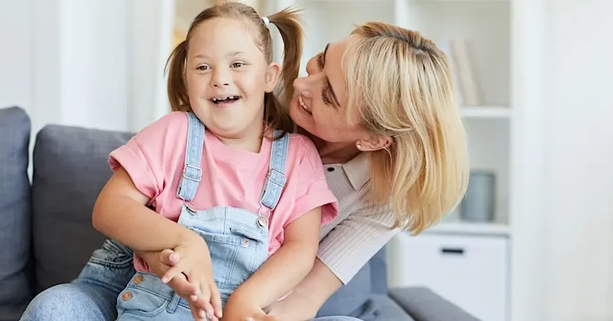Woman sitting with a child in her lap. Woman has got her face up by the girls ear and the girl is smiling.