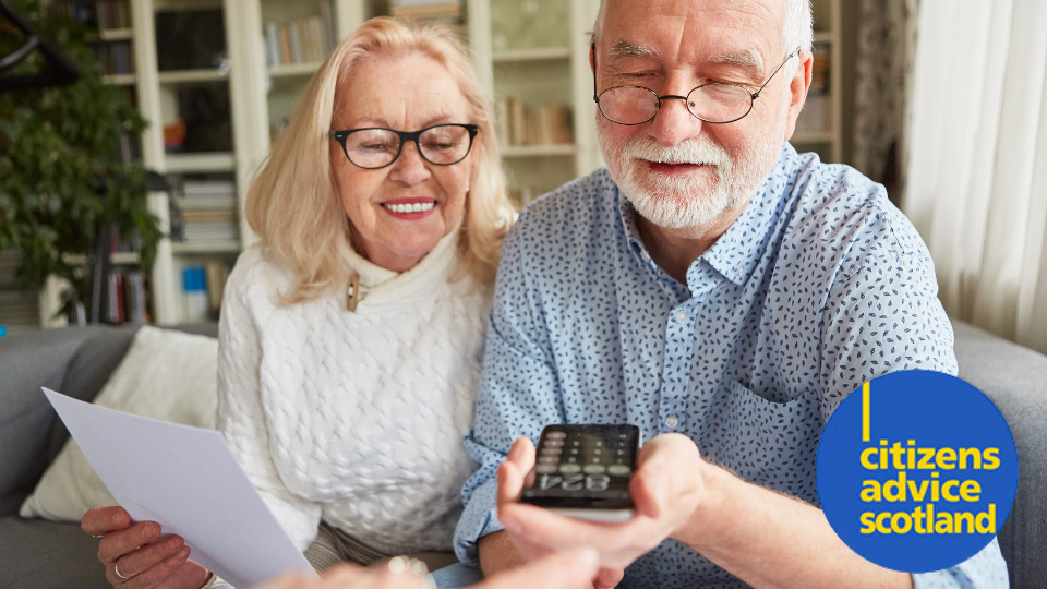 Older man and woman sitting together smiling at a calculator in the man's hand.