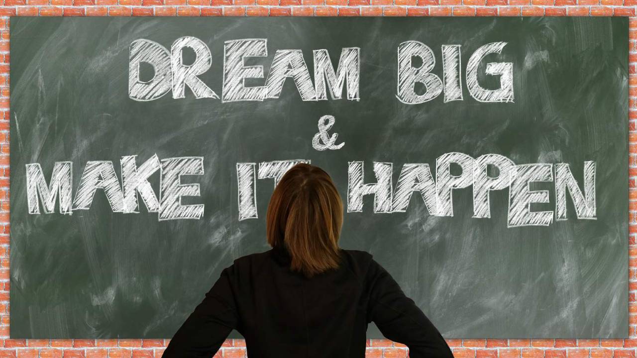 Person with long brown hair and a black top looks at a blackboard that reads 'Dream big & Make it Happen'