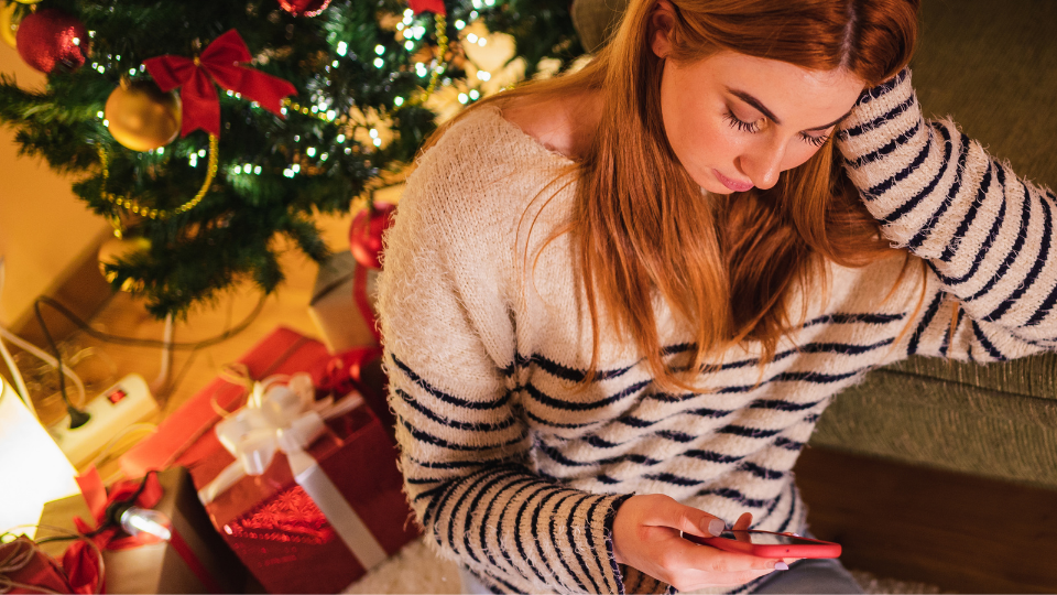 Downward view of a woman looking at a mobile phone, sitting crosslegged on living room floor with her elbow leaning on a sofa and a christmas tree behind her.