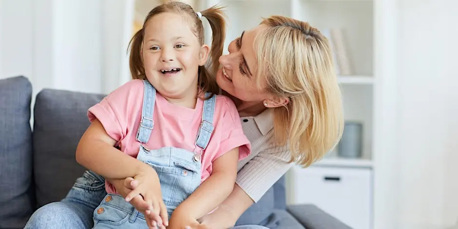 Woman sitting with a child in her lap. Woman has got her face up by the girls ear and the girl is smiling.