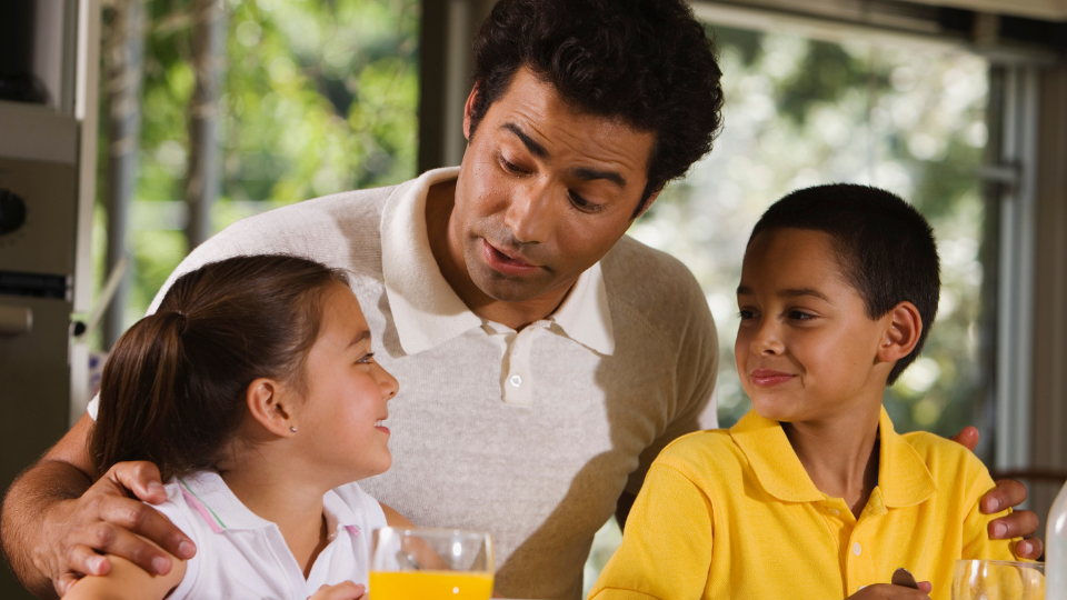 A man is sitting with his arms around a boy and a girl. The man and the boy are looking at the girl who is speaking.