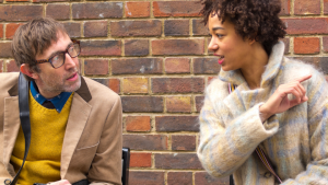 A man and a woman sitting outside against a brick wall backdrop looking at each other while having a conversation. The woman is pointing to something off camera.