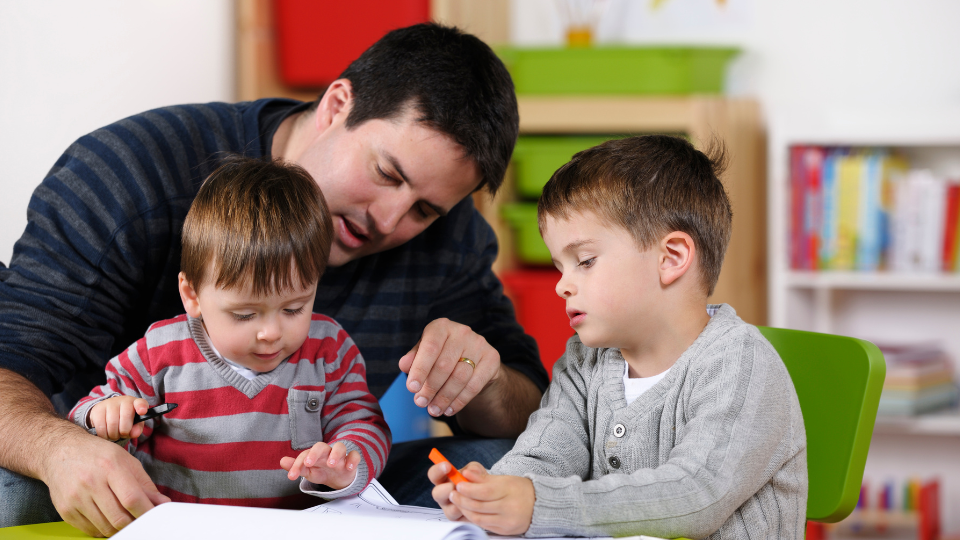 Image of a man sittting with a toddler sitting on his lap and a young boy sitting next to him. The group are doing a creative activity.