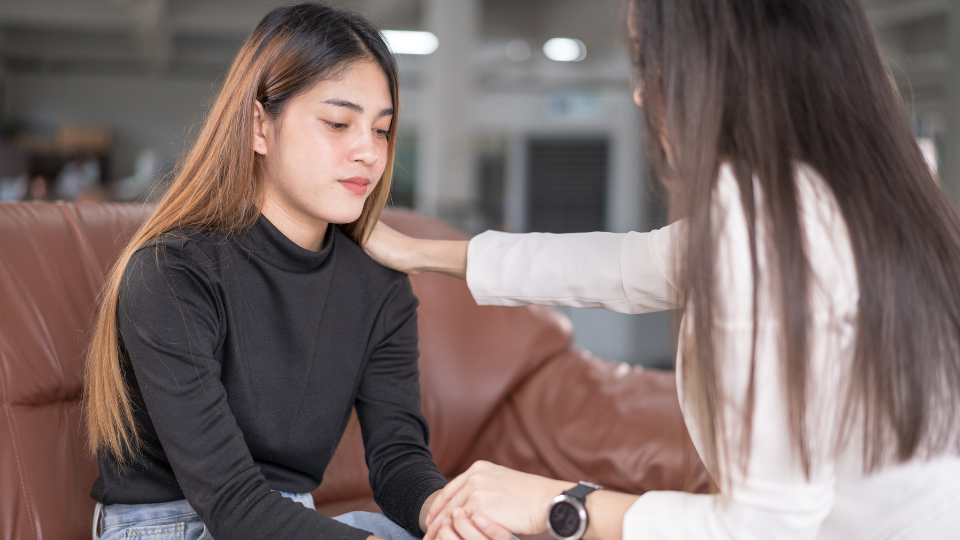 Young adult women sitting on a sofa looking down and sad. A woman sits in front with hand on young womans shoulder and holding her other hand in comfort.